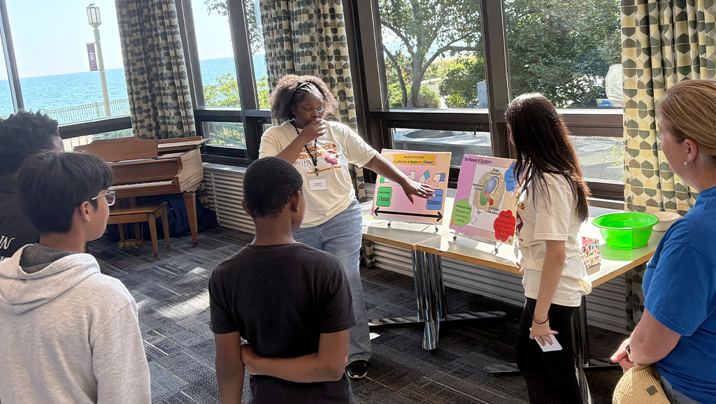 Two students present colorful posters about the brain to a small group of middle and high school students during a hands-on learning event in a lakeside campus classroom.